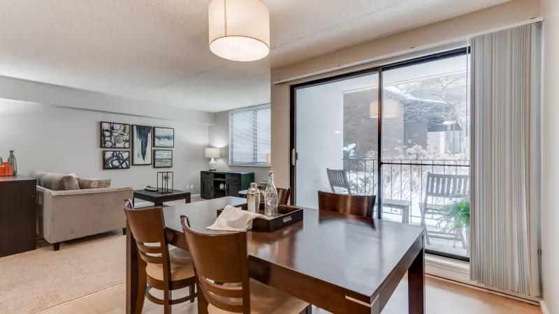 Bright dining area with a wooden table, four chairs, and a view into a living room and balcony.