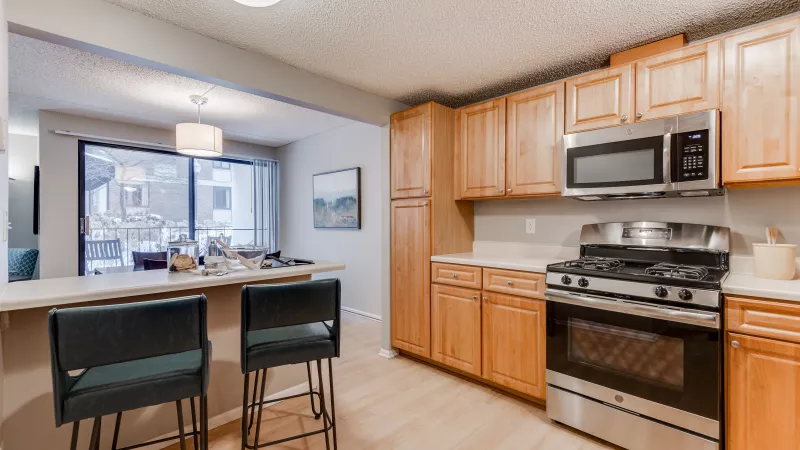 Modern kitchen with light wood cabinets, stainless steel appliances, breakfast bar, and two dark stools.