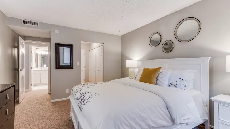Bright bedroom with a white bed, yellow pillow, three mirrors, and a view into a bathroom.