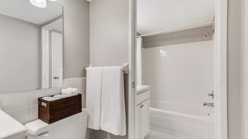 Clean, bright white bathroom with a toilet, a wooden basket of towels, a towel bar, and a white bathtub with a shower.