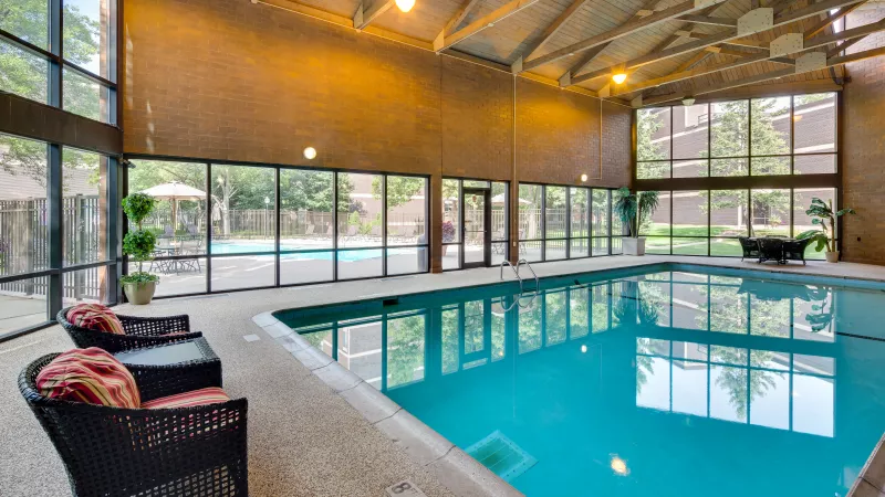 Indoor swimming pool with large windows, bright blue water, and two wicker chairs in the foreground.