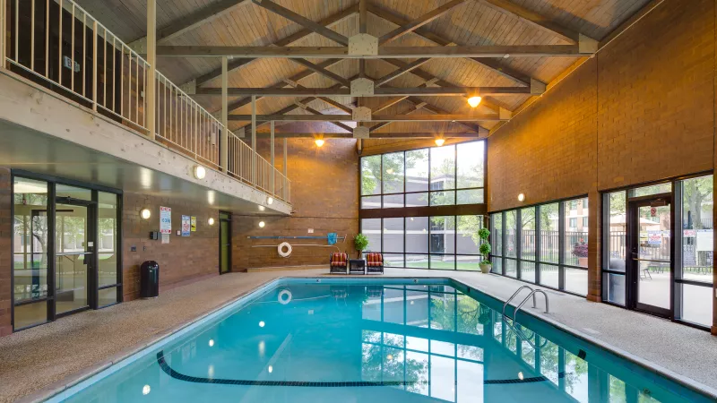 Indoor swimming pool with a high wood-beam ceiling, brick walls, large windows, and an upper balcony.