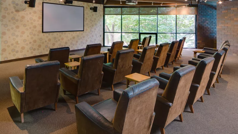 Empty screening room with rows of brown leather armchairs, a large projection screen, and windows showing green trees.