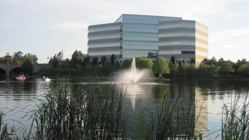 Modern glass office building across a serene lake with a central fountain. Reeds frame the foreground; people on pedal boats.