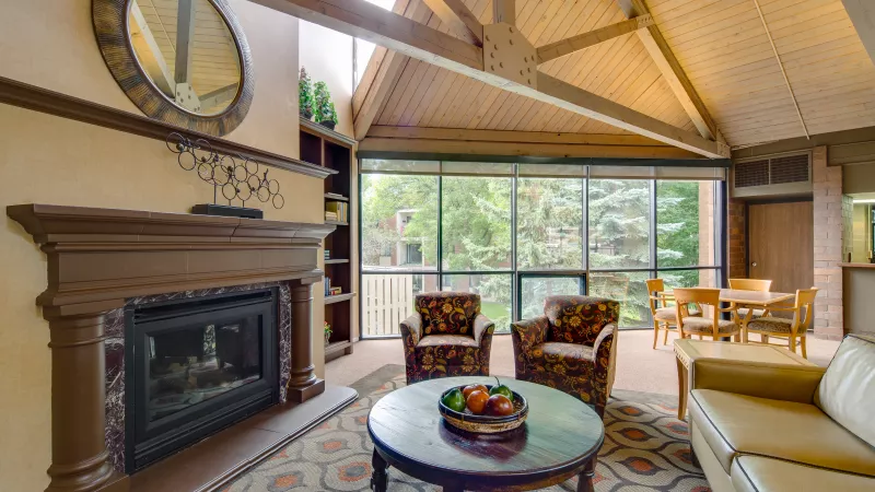 Spacious living room with a high beamed ceiling, fireplace, patterned armchairs, and a large window viewing trees outside.