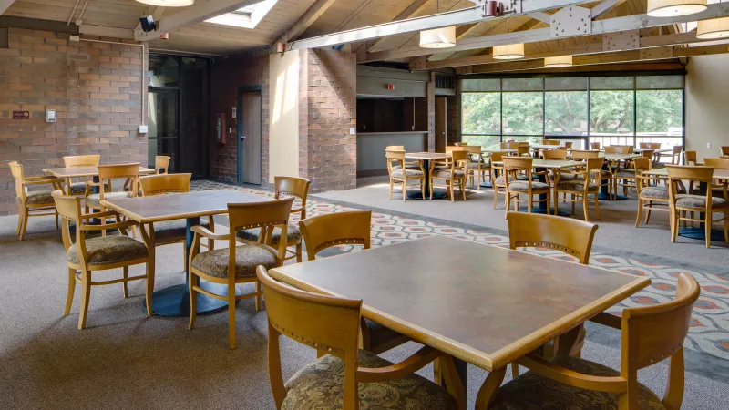 Empty dining hall with many square tables, wood-framed chairs, a vaulted ceiling, and large windows.