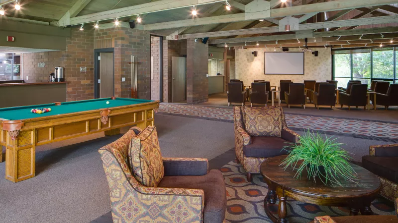 Recreation lounge with a billiards table, patterned armchairs, a bar, and a raised presentation area with screen and chairs.