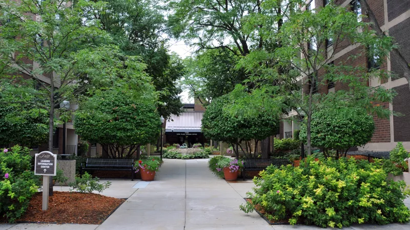 Paved urban courtyard lined with lush green trees, shrubs, benches, and colorful potted flowers.