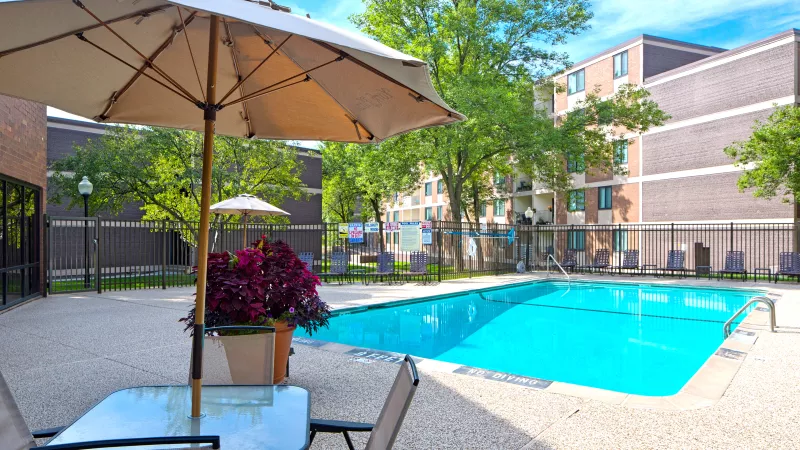 Sunny outdoor swimming pool with a patio table, chairs, and beige umbrella. Apartment buildings and green trees in the background.