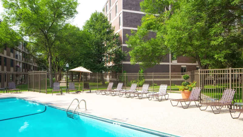 Bright outdoor swimming pool area with numerous lounge chairs, green trees, and a brick apartment building.