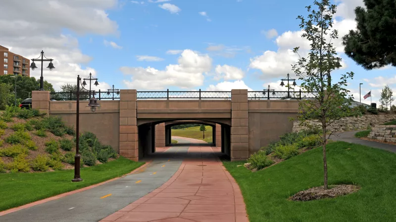 Red and grey paved paths merge, leading into a concrete underpass beneath a bridge on a sunny day.