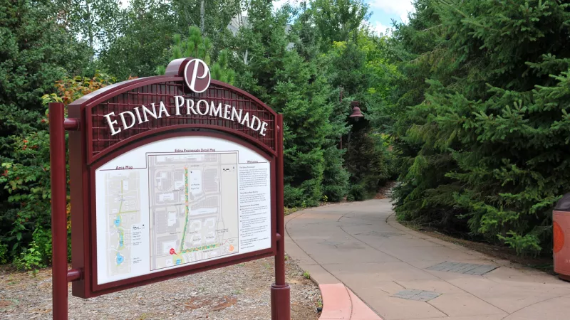 Brown Edina Promenade sign with map, beside a winding paved path leading into dense green trees.