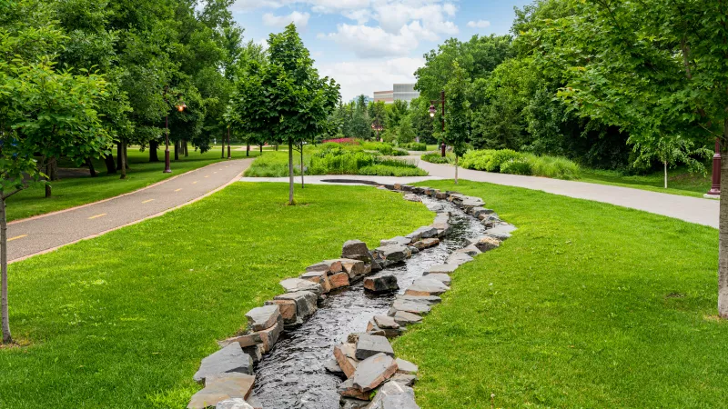 Winding rock-lined stream flows through a sunny green park with walking paths and lush trees under a bright sky.