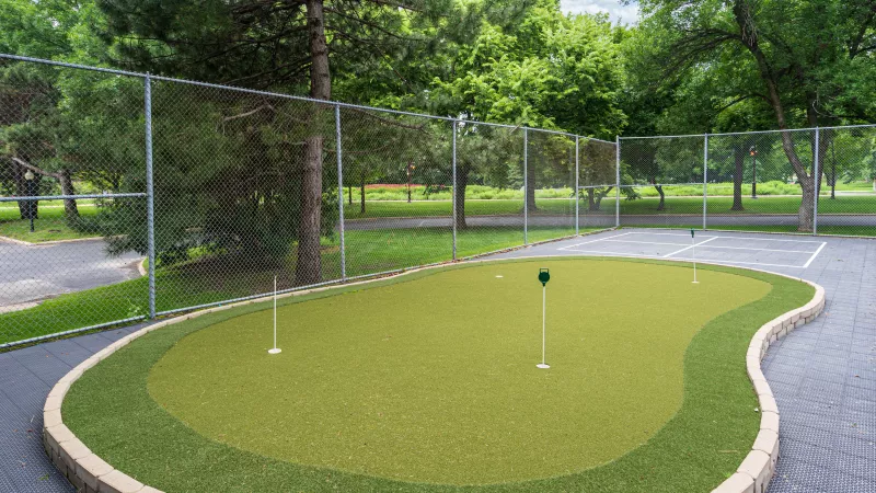 Outdoor putting green with green artificial turf and three flags, fenced, surrounded by trees.