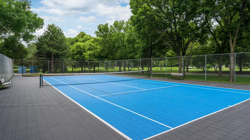 Blue and gray outdoor tennis court surrounded by green trees and a chain-link fence under a partly cloudy sky.