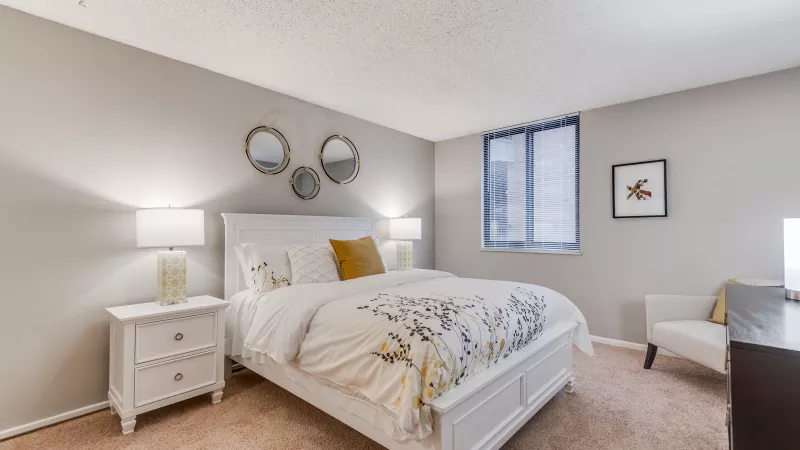 Bright bedroom with a white bed, yellow and gray floral bedding, two nightstands with lamps, and decorative mirrors.