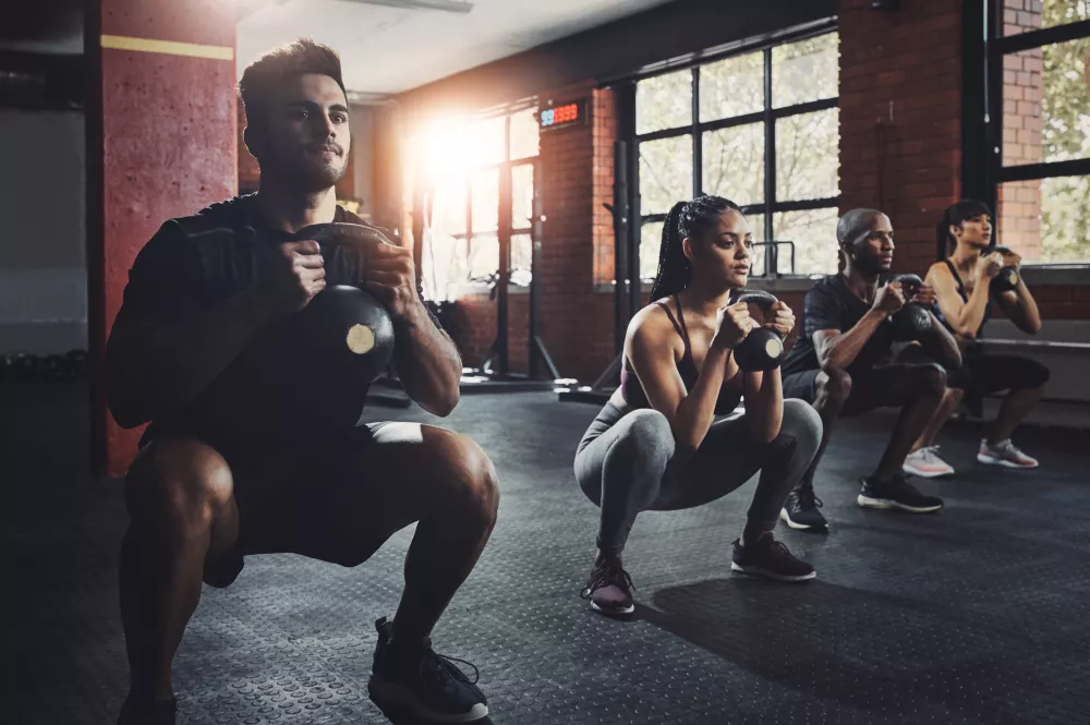 Four people doing kettlebell squats in a sunlit gym with red walls.