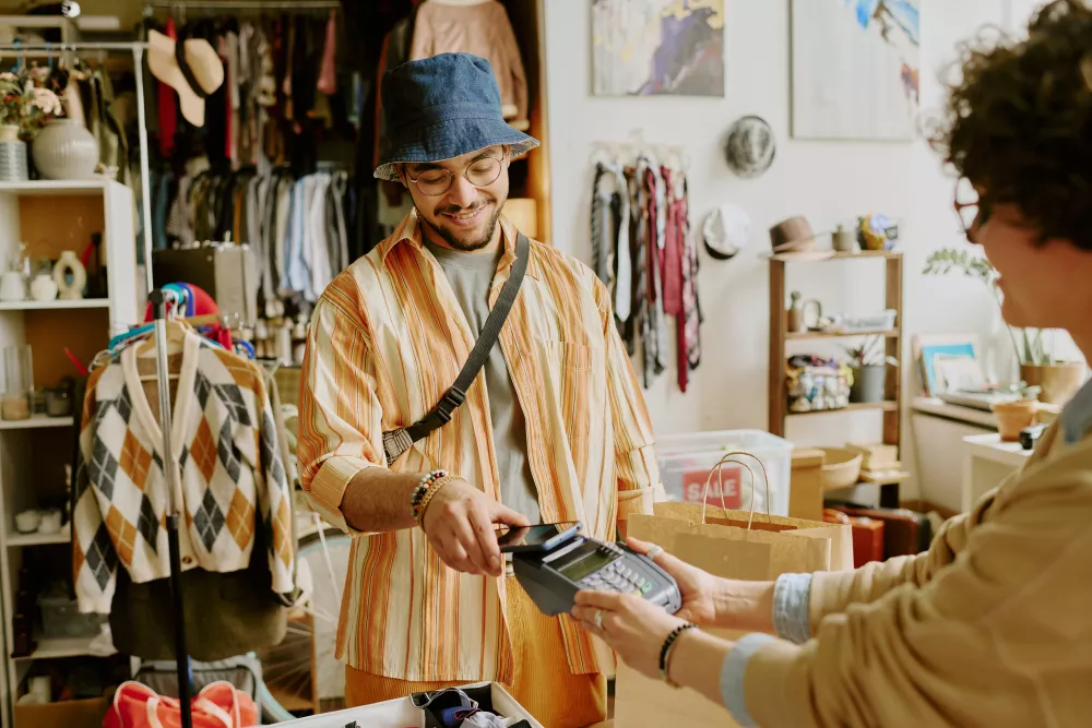 Smiling man in a bucket hat paying with a card at a clothing store, cashier holds the payment terminal.
