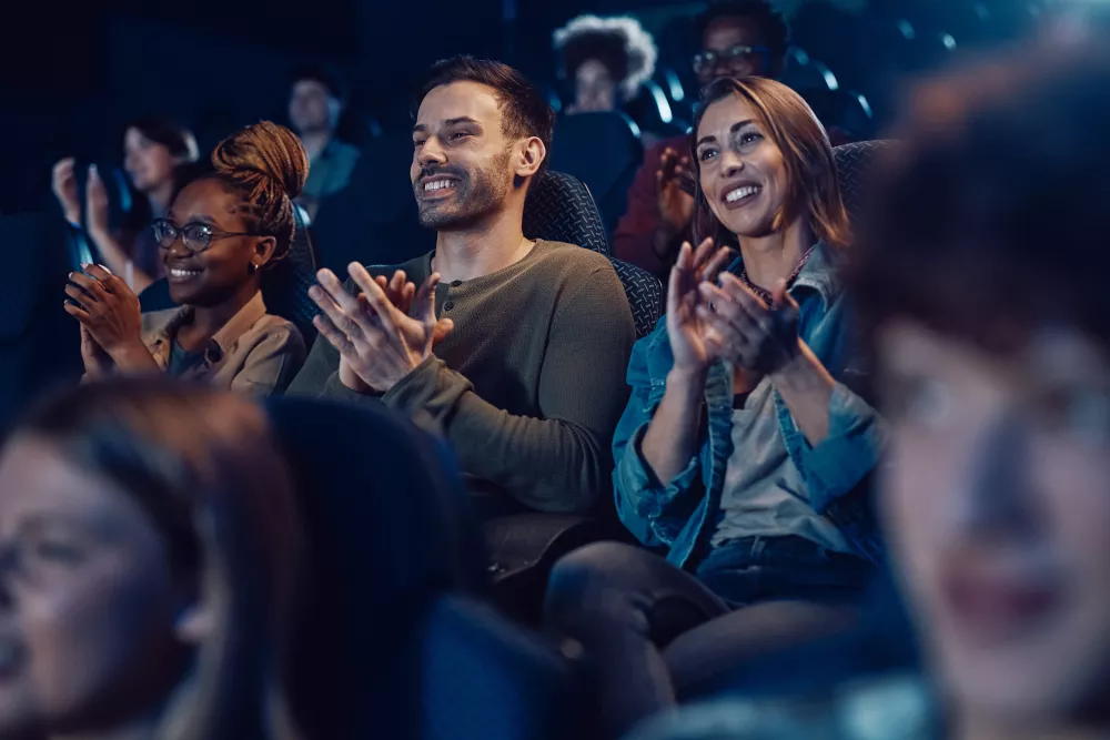 Happy diverse audience in a dark movie theater, clapping and smiling.