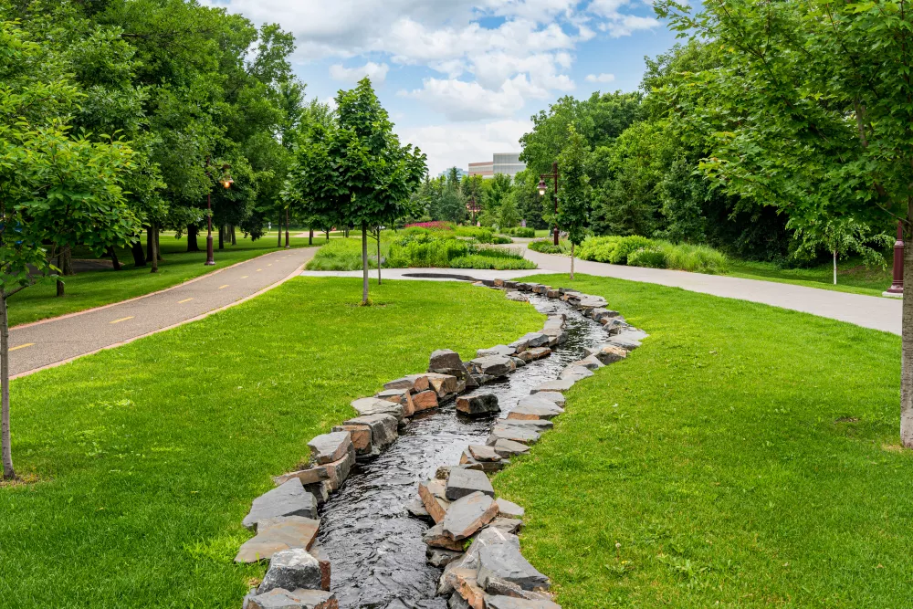 Winding rock-lined stream flows through a sunny green park with walking paths and lush trees under a bright sky.